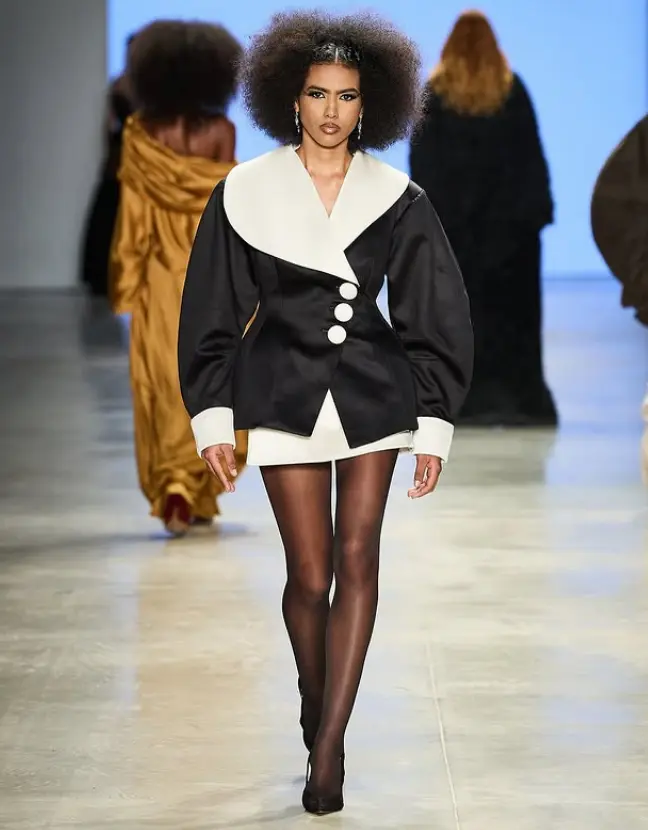 Modelo desfilando com um visual inspirado nos anos 40, com um casaco preto e branco que remete à elegância de Hollywood.