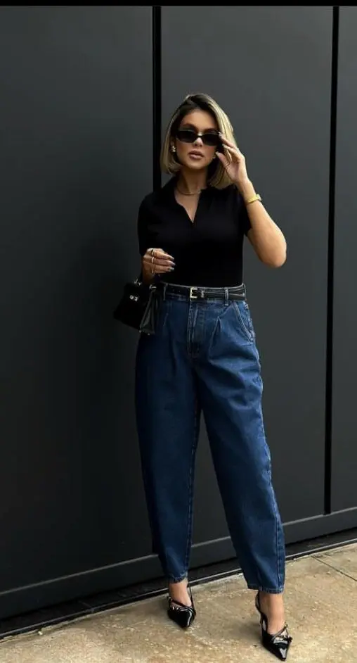 Mulher com look elegante de calça jeans escura e camisa polo preta, usando óculos de sol e salto alto.