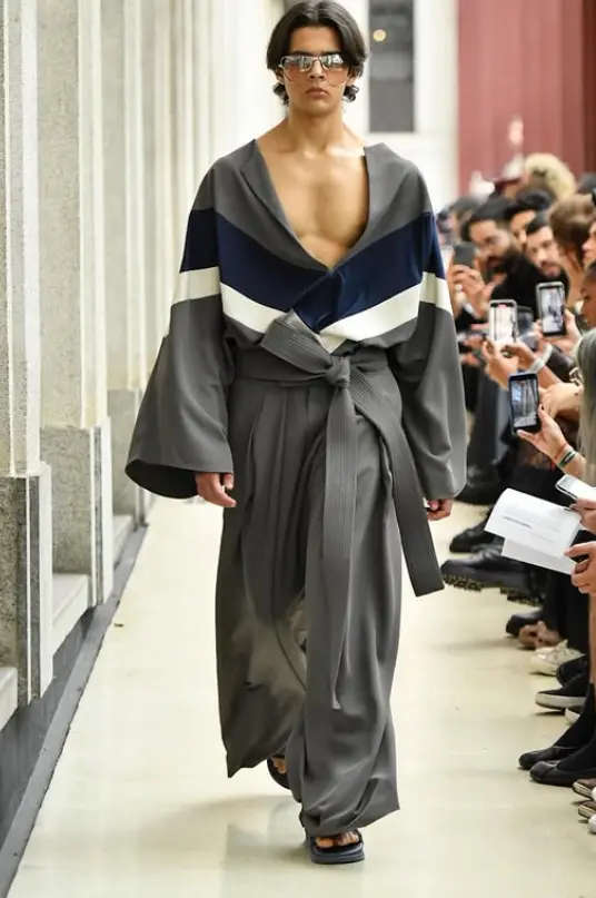 Homem desfilando na SPFW N60 com um look de roupa de estilo japonês, com um nó de destaque na parte da cintura.