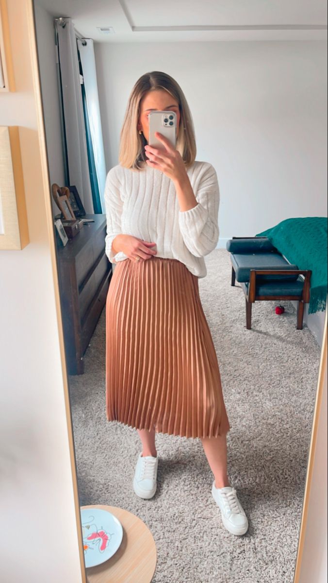 Mulher em um espelho, usando um suéter branco de tricô, saia plissada e tênis branco, mostrando um look confortável e moderno.