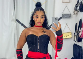 Mulher com fantasia de ninja, com vestido preto, fita vermelha e acessórios, posando para foto.