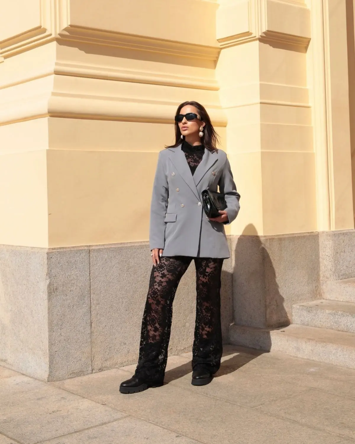 Mulher estilosa com blazer cinza e calça de renda preta, completando o look com acessórios minimalistas.