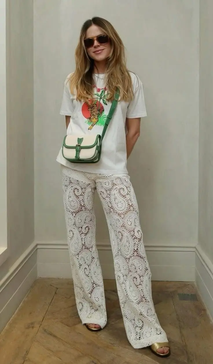 Mulher usando calças de renda branca e camiseta estampada com um tigre, bolsa verde e óculos escuros, posando em ambiente neutro.