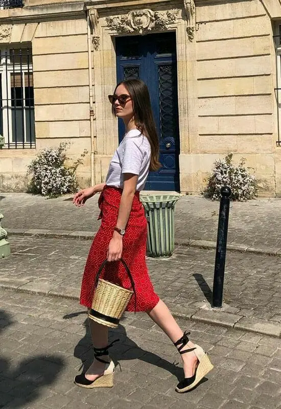 Mulher caminhando com saia midi vermelha, sandálias anabela e bolsa de palha em uma rua clássica.