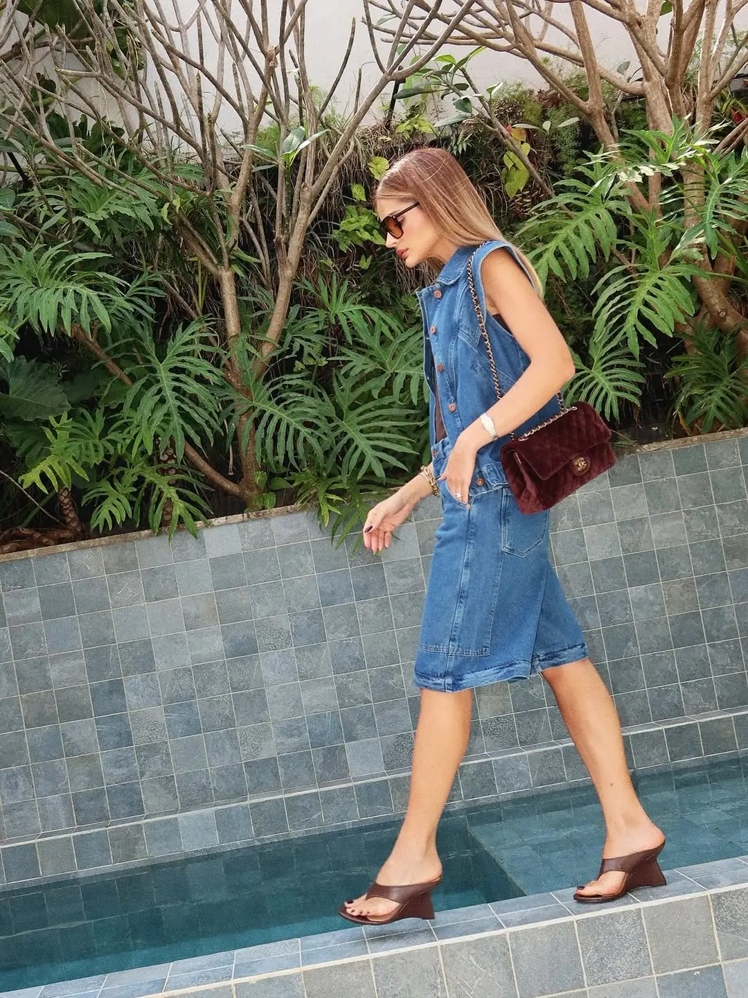 Mulher caminhando ao lado de uma piscina vestindo conjunto jeans com bermuda comprida, bolsa de veludo e salto anabela.