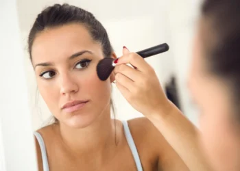 Mulher aplicando blush em sua pele seca e desidratada diante de um espelho.