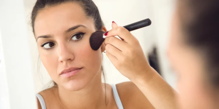 Mulher aplicando blush em sua pele seca e desidratada diante de um espelho.