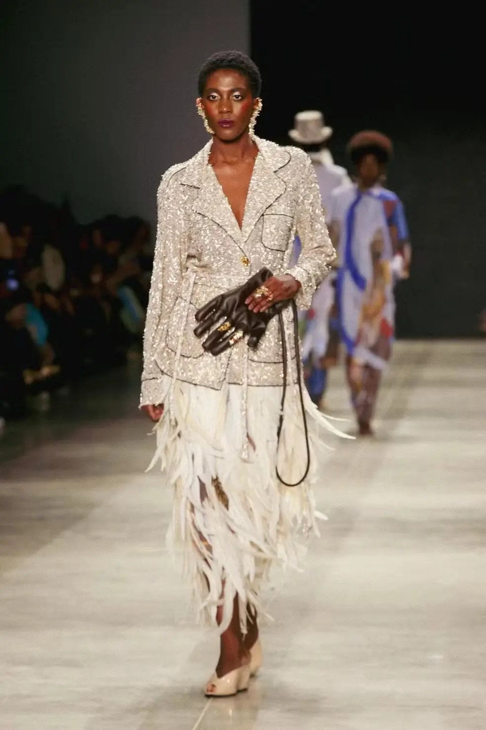 Modelo desfilando no SPFW com blusa de paetê e saia de penas, carregando uma bolsa em forma de luva.