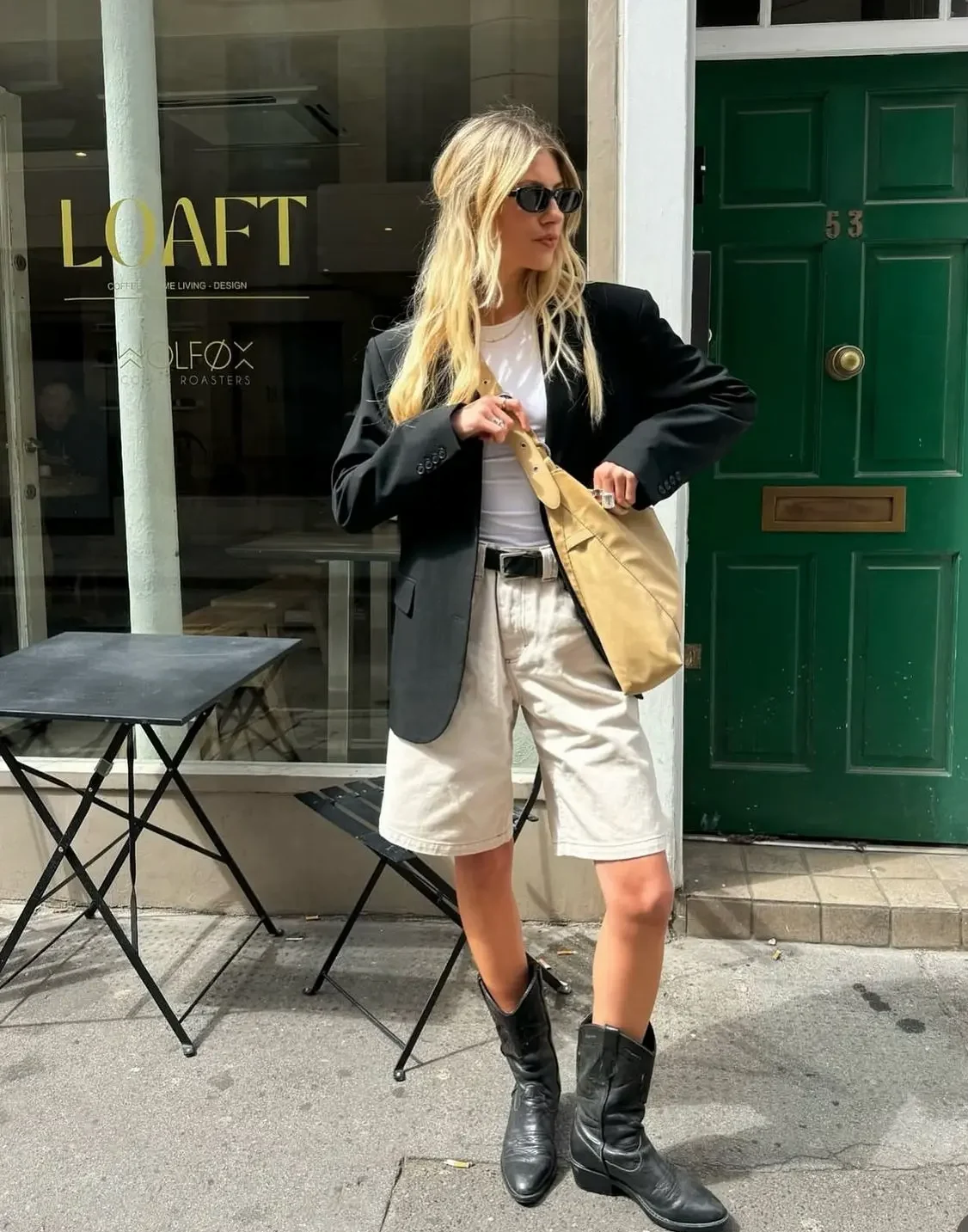 Mulher com blazer preto, camiseta branca e bermuda clara, usando botas de cowboy pretas.