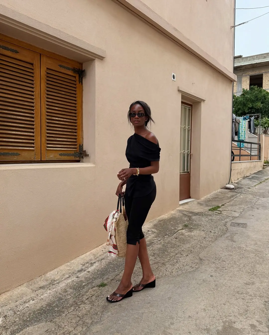 Mulher elegante caminhando em uma rua com calça capri preta, blusa ombro a ombro e sandália anabela, transmitindo um estilo moderno e retrô.