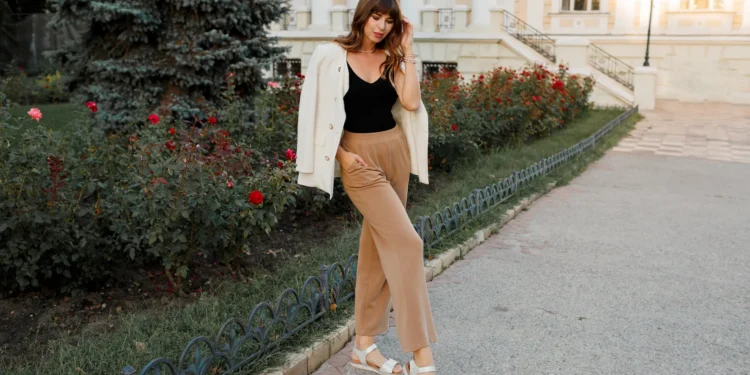 Mulher posando na rua com jaqueta branca, calça confortável e sandálias, em frente a um jardim de rosas.