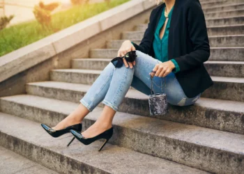 Mulher sentada em escadaria usando calça jeans justa com salto alto preto e blazer elegante.
