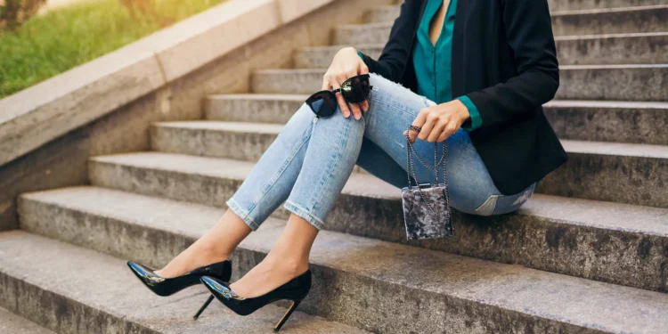 Mulher sentada em escadaria usando calça jeans justa com salto alto preto e blazer elegante.