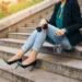Mulher sentada em escadaria usando calça jeans justa com salto alto preto e blazer elegante.