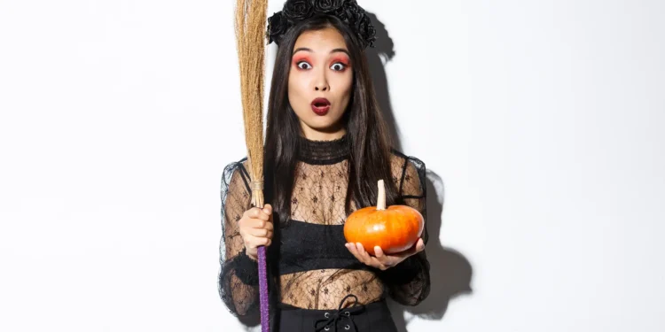 Mulher com expressão surpresa vestida de bruxa segurando abóbora e vassoura para Halloween.