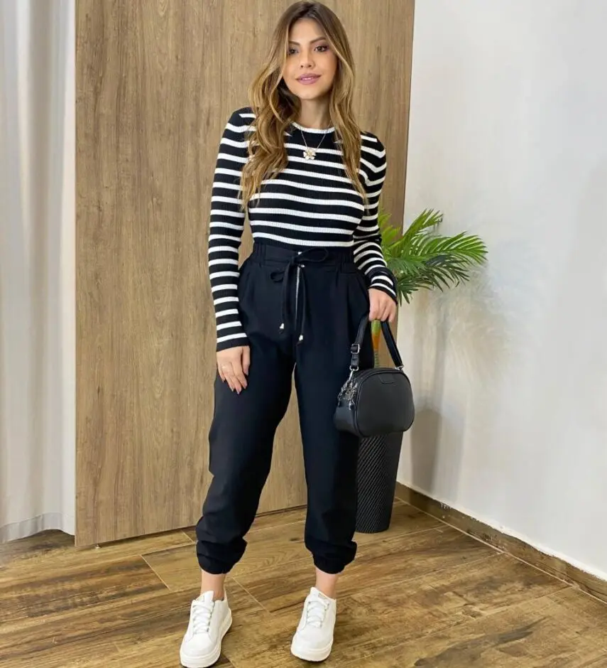 Mulher usando calça jogger de viscose preta com blusa listrada preta e branca, tênis branco e bolsa preta.