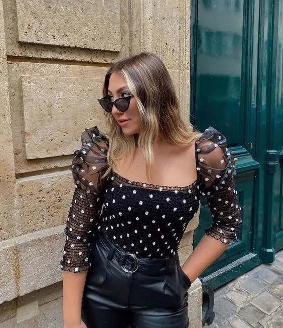 Mulher usando blusa de poá de lastex preta e calça de couro preta, com óculos escuros, posando na rua.