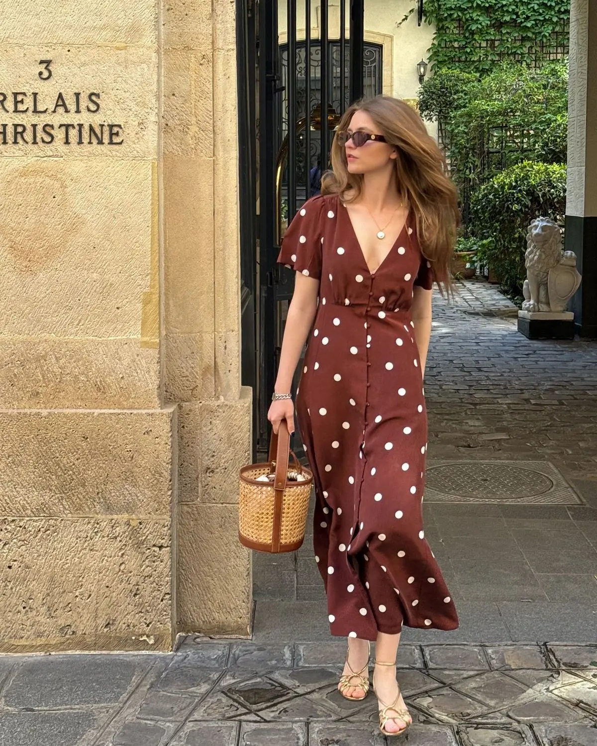 Mulher usando vestido midi de poá marrom com acessórios e óculos de sol, caminhando em uma rua parisiense.