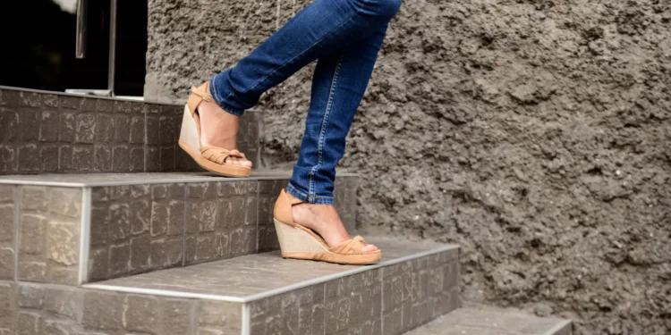 Salto anabela está em alta novamente! Veja como usar nos seus looks 1 Mulher usando sandálias anabela bege enquanto sobe escadas de pedra.