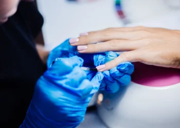 Profissional aplicando esmalte de gel em unha de cliente com luvas azuis.