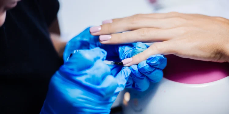 Profissional aplicando esmalte de gel em unha de cliente com luvas azuis.