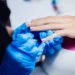 Unhas de gel: 10 dicas essenciais para manter suas unhas bonitas e saudáveis 17 Profissional aplicando esmalte de gel em unha de cliente com luvas azuis.