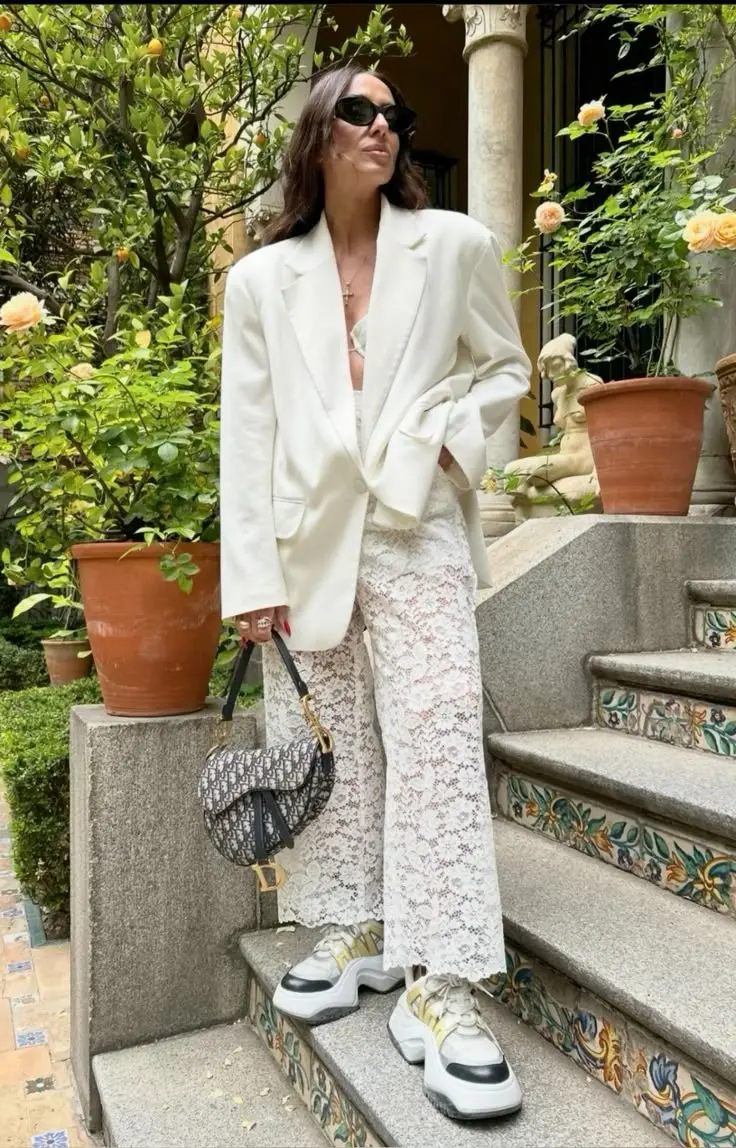 Mulher usando blazer branco oversized com calça de renda e tênis, com uma bolsa Dior, em um ambiente ao ar livre.