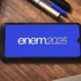 Tela de celular exibindo o logotipo do ENEM 2025 sobre uma mesa com cadernos e canetas.