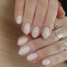 Unhas curtas com esmalte nude e acabamento brilhante, com anéis dourados delicados, ideal para um visual discreto e sofisticado.