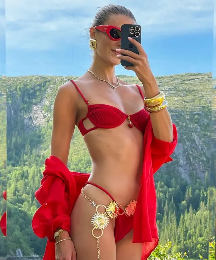 Verão 2026: Confira 5 cores de biquíni que serão destaque na temporada 3 Mulher fotografando-se com biquíni vermelho tomate e acessórios dourados diante de paisagem montanhosa.