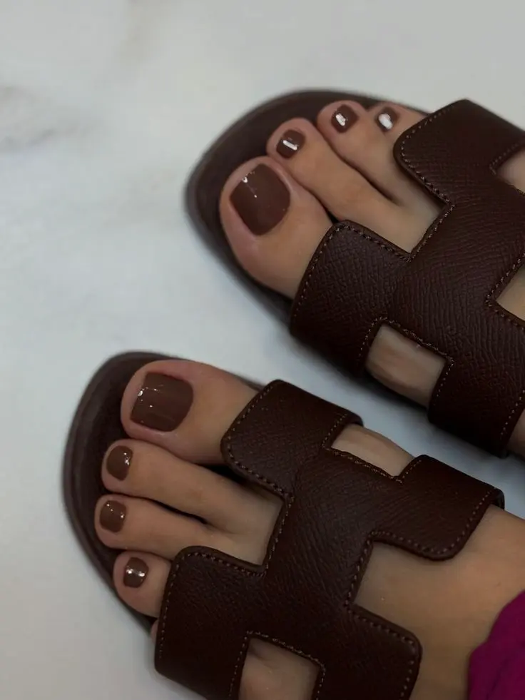 Cores de esmalte para os pés que serão tendência no verão 2026; renove seu visual 3 Pés com esmalte marrom terroso em sandálias marrons.